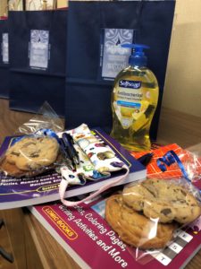 A close-up of a puzzle book, masks, a bag of cookies tied with a blue ribbon, Softsoap Soap, and two pens and navy gift bags in the background