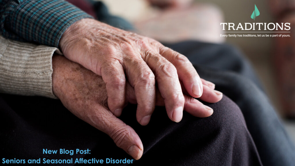 A close-up of two elderly hands, one hand is laying on top and the other than is resting on their knee. Traditions Logo on the top right corner. New Blog Post: Seniors and Seasonal Affective Disorder in blue text at the bottom left corner.