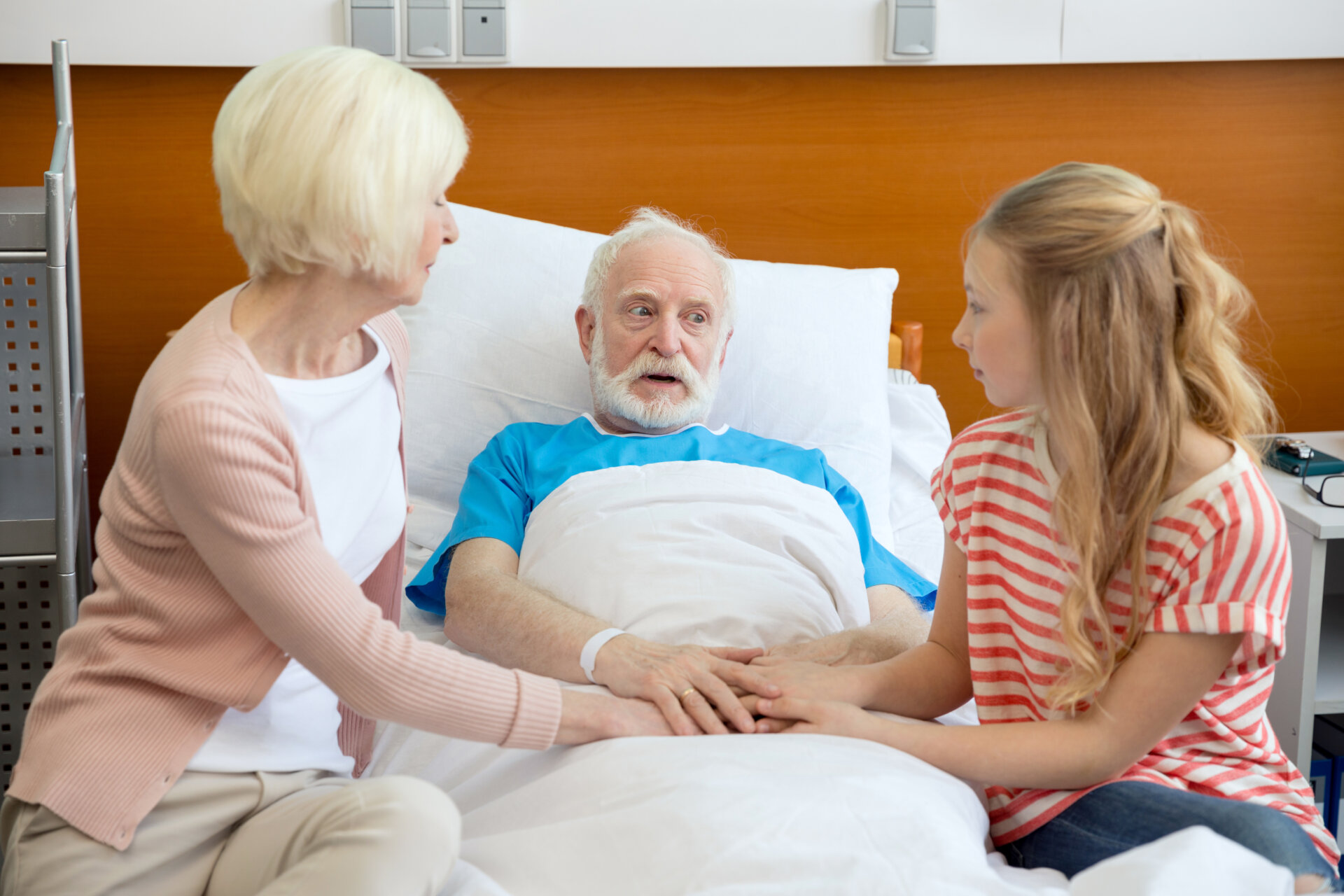 A young girl and an elderly woman holding an elderly man's hands who is lying on the bed