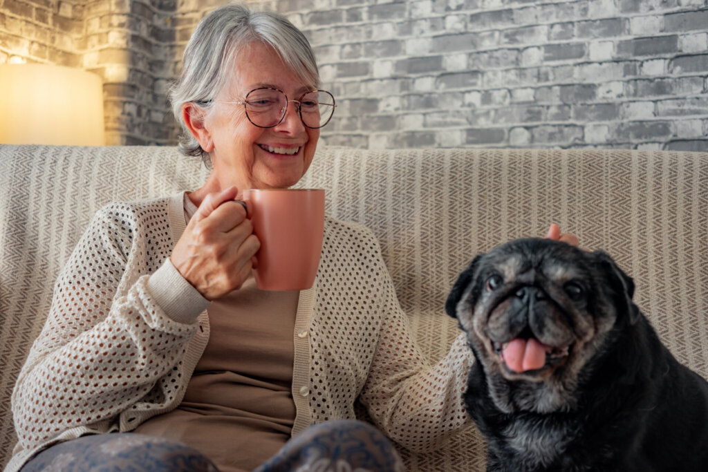 Senior happy woman sitting on sofa with her best friend, an old black pug dog. Elderly lady relax with a coffee cup in good company