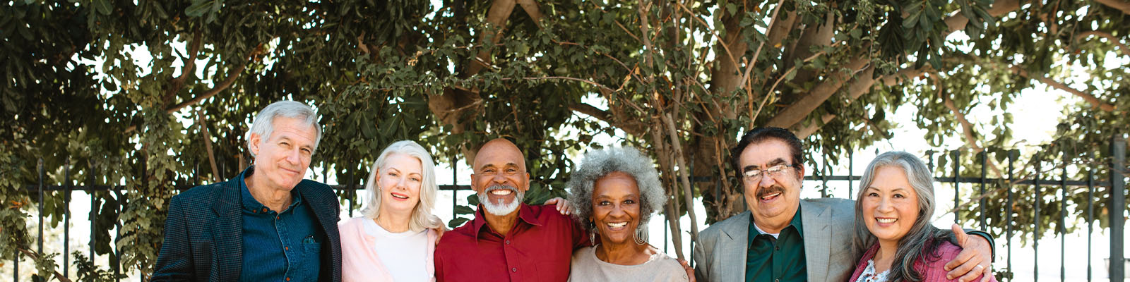A group of senior men and women standing outside together in front of a large tree with their arms around each other