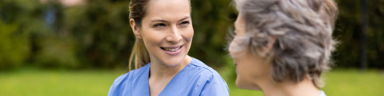 A senior woman walking outdoors with her female nursing assistant as they smile at each other
