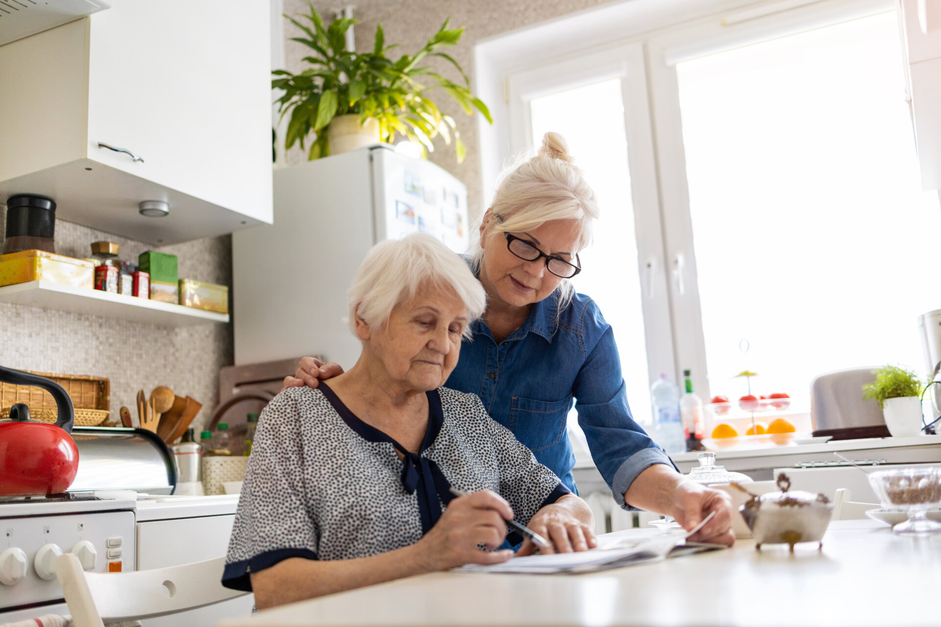 This is a close-up of a younger and older woman around a kitchen table. The younger woman is leaning down and holding onto the older woman's back, looking down at the paperwork.