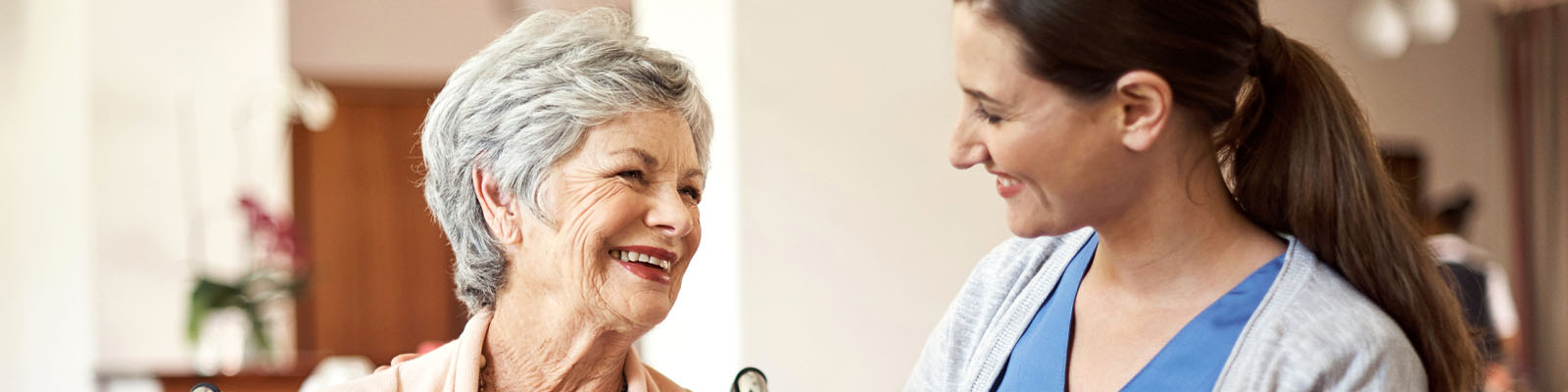 A senior woman sitting in a wheelchair looking over at her nursing assistant while smiling