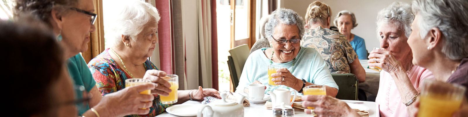 A group of senior women sitting together at a table in a dining area having breakfast and drinking orange juice
