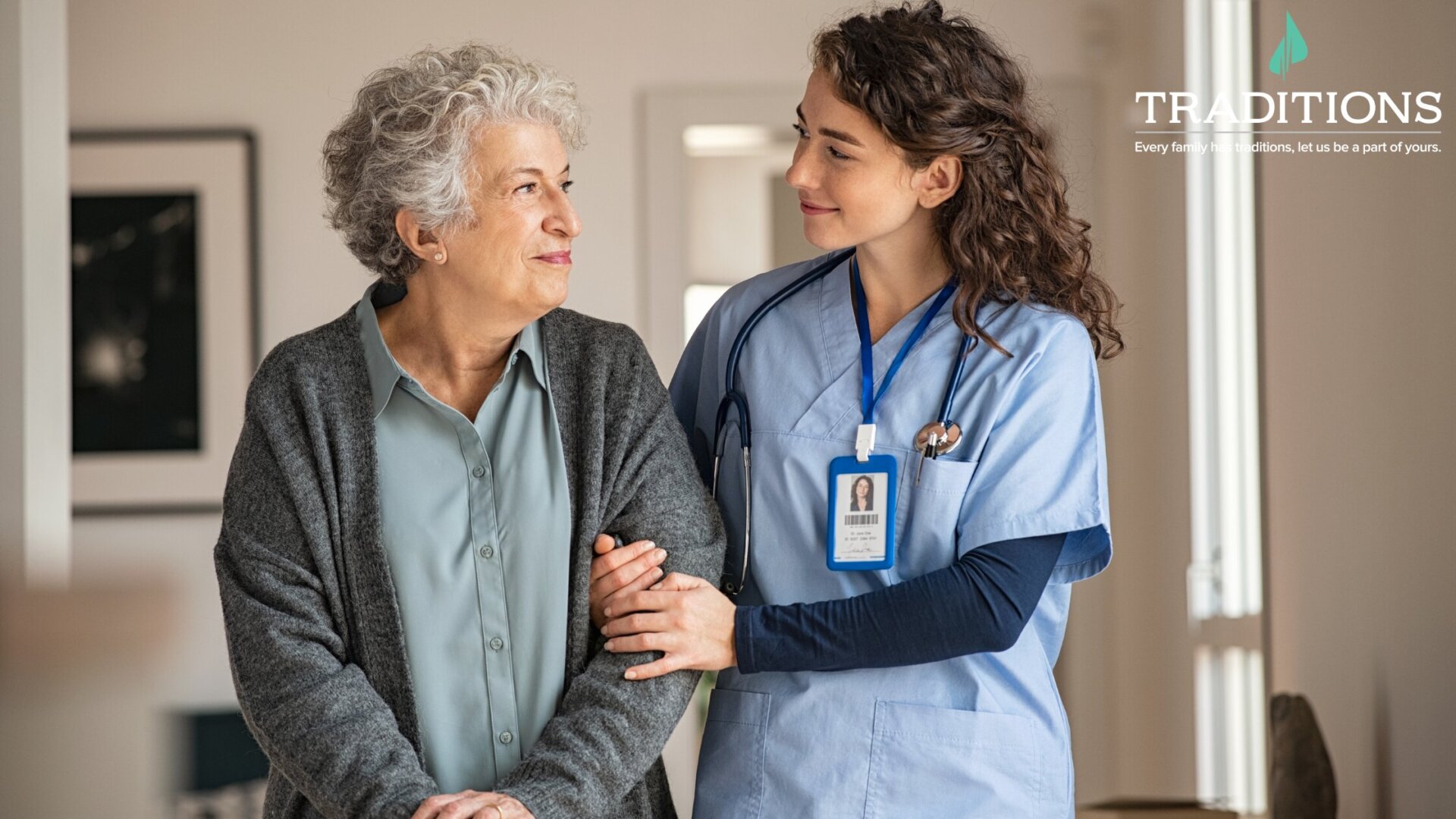 A female caregiver holds an older woman's arm while walking down a hall with the Traditions logo at the top right corner.