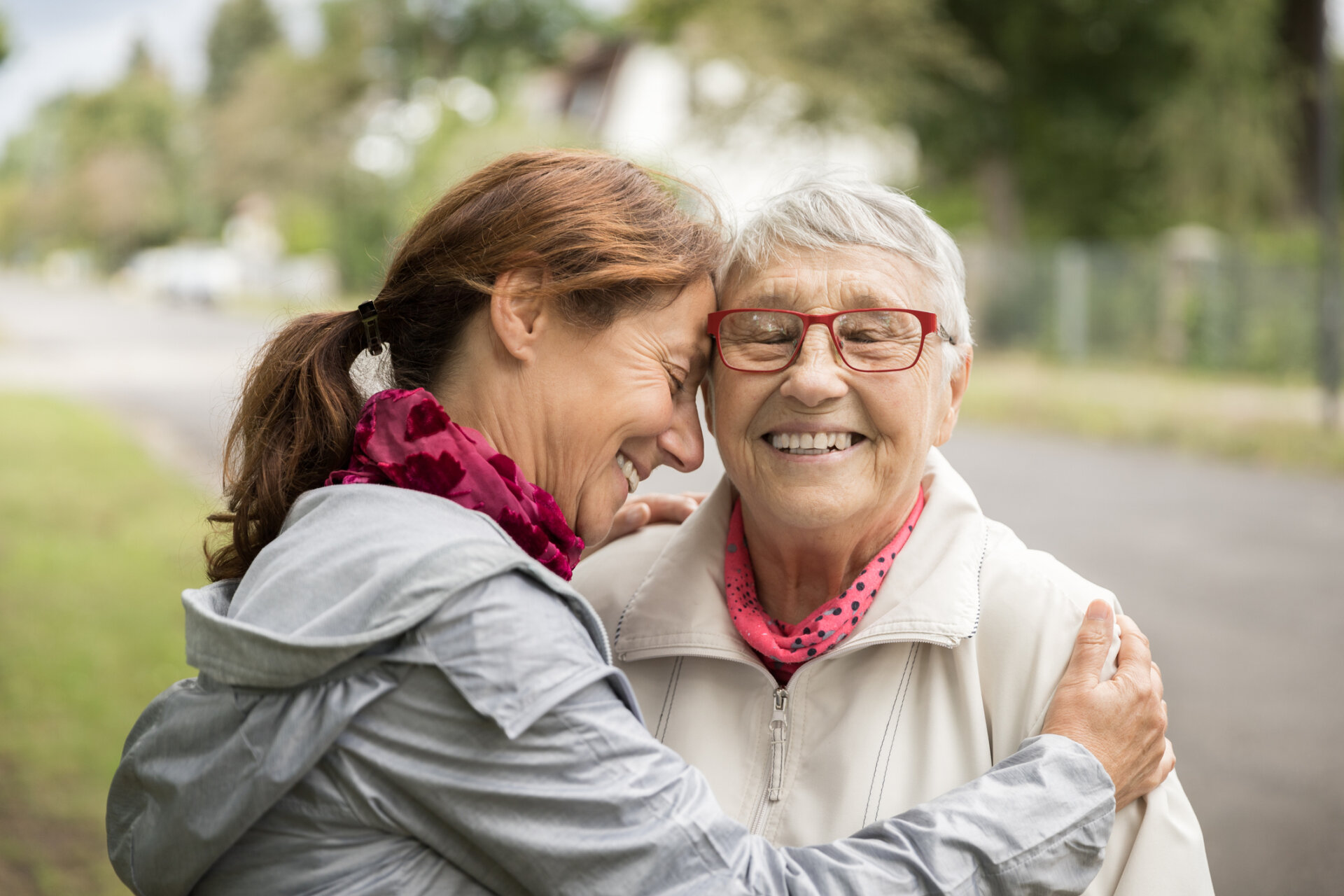 Two women, both wearing raincoats outside, hugging each other and smiling.
