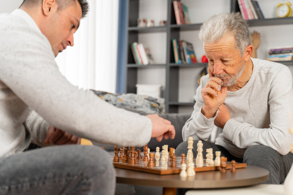 Male senior memory care resident playing chess at a table in his memory care apartment at Traditions at Camargo with his son