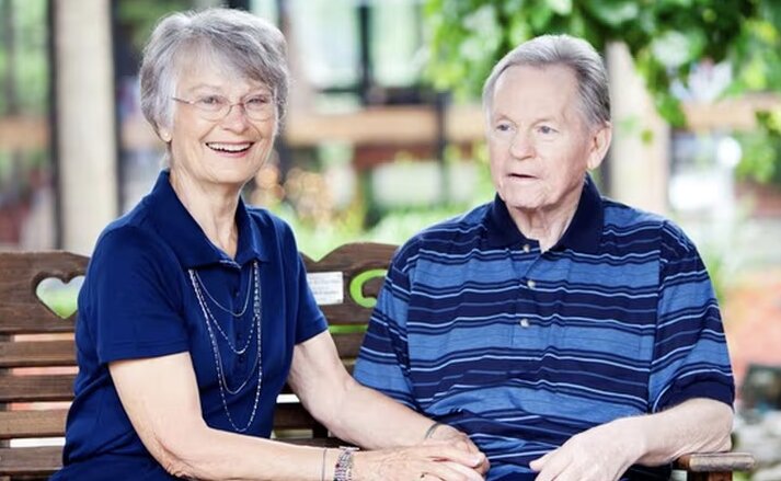 A couple sitting outside on a bench smiling. The woman wears a blue top and glasses, and the man wears a striped blue polo. Traditions at Camargo